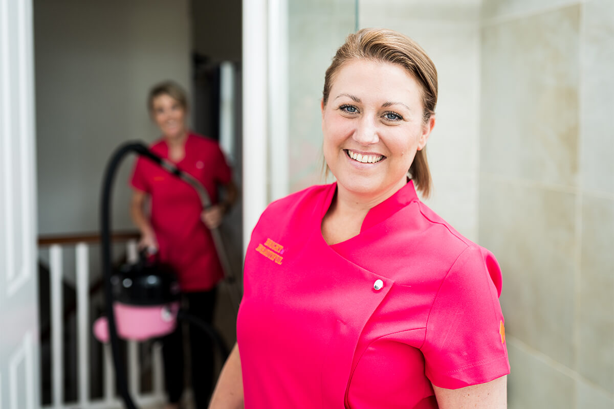 friendly housekeeper smiles at camera whilst colleague hoovers in the background